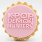 Round pink fondant cookie embossed with the text “KPOP DEMON HUNTERS” in chunky letters.